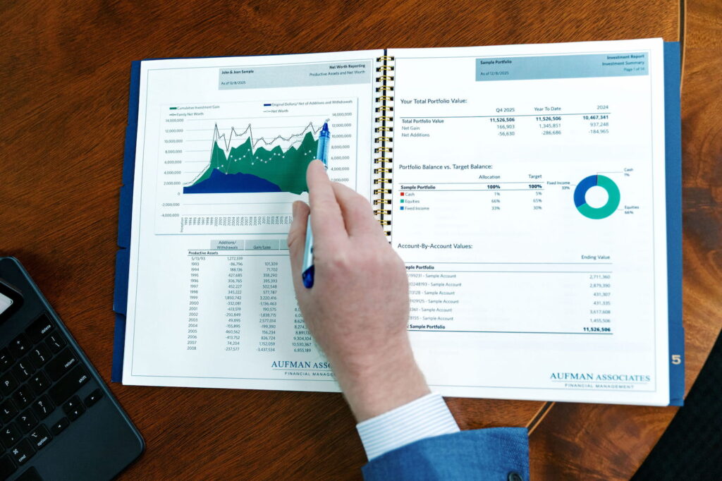 a printed client date spreadsheet booklet lays open on a dark wood table while an associate points to data with a pen in hand