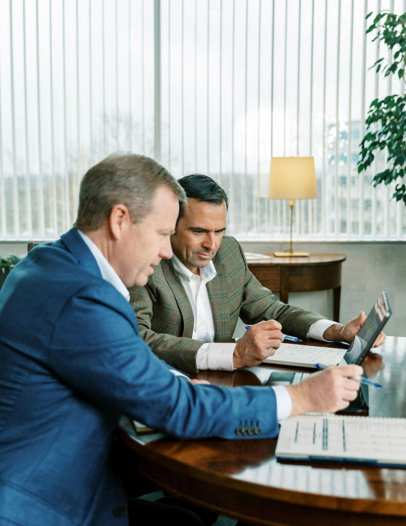 two Aufman Associates sit at a table looking at a laptop screen while going over spreadsheets and data with a client