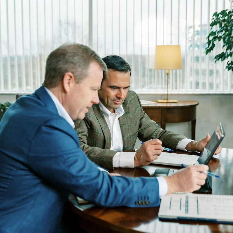 two Aufman Associates sit at a table looking at a laptop screen while going over spreadsheets and data with a client