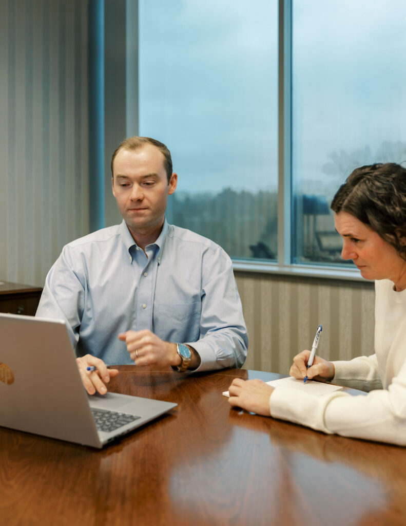 two Aufman Associates sit at a table looking at a laptop screen while going over spreadsheets and data with a client